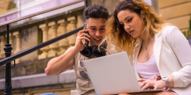 Young entrepreneurs doing teamwork. Working on the internet with the computer, boy with music headphones and girl in white suit / Envato Dos jóvenes mirando una app de finanzas