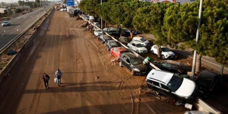 Dos personas caminan por la V-31 en Sedaví (Valencia) junto a decenas de coches apilados contra el quitamiedos, este jueves. / EL PAIS Claudio Álvarezvarez Dos personas caminan por la V-31 en Sedaví (Valencia) junto a decenas de coches apilados contra el quitamiedos, este jueves.