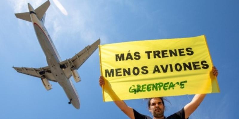 Cartel en el que se lee 'Más trenes, menos aviones', con un avión sobrevolando de fondo | Foto de Greenpeace Un activista de Greenpeace sostiene un cartel en el que se lee 'Más trenes, menos aviones', con un avión sobrevolando de fondo