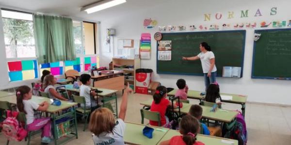 Aula de colegio en la que la profesora explica junto a la pizarra de color verde