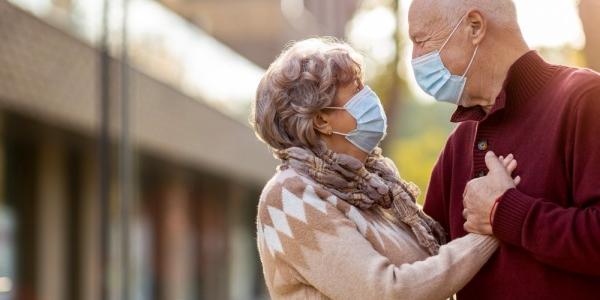 Una pareja de personas mayores con mascarilla se miran abrazados 