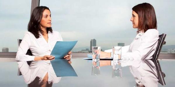 Dos mujeres separada por una mesa durante una entrevista de trabajo. Ambas visten con ropa clara /casacochecurro.com