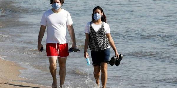 Un chico y una chica pasean por la playa con la mascarilla en el rostro 