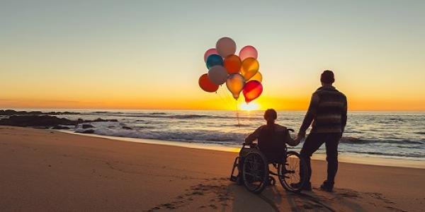 Persona en silla de ruedas junto a un acompañante disfrutan de un atardecer en el mar 